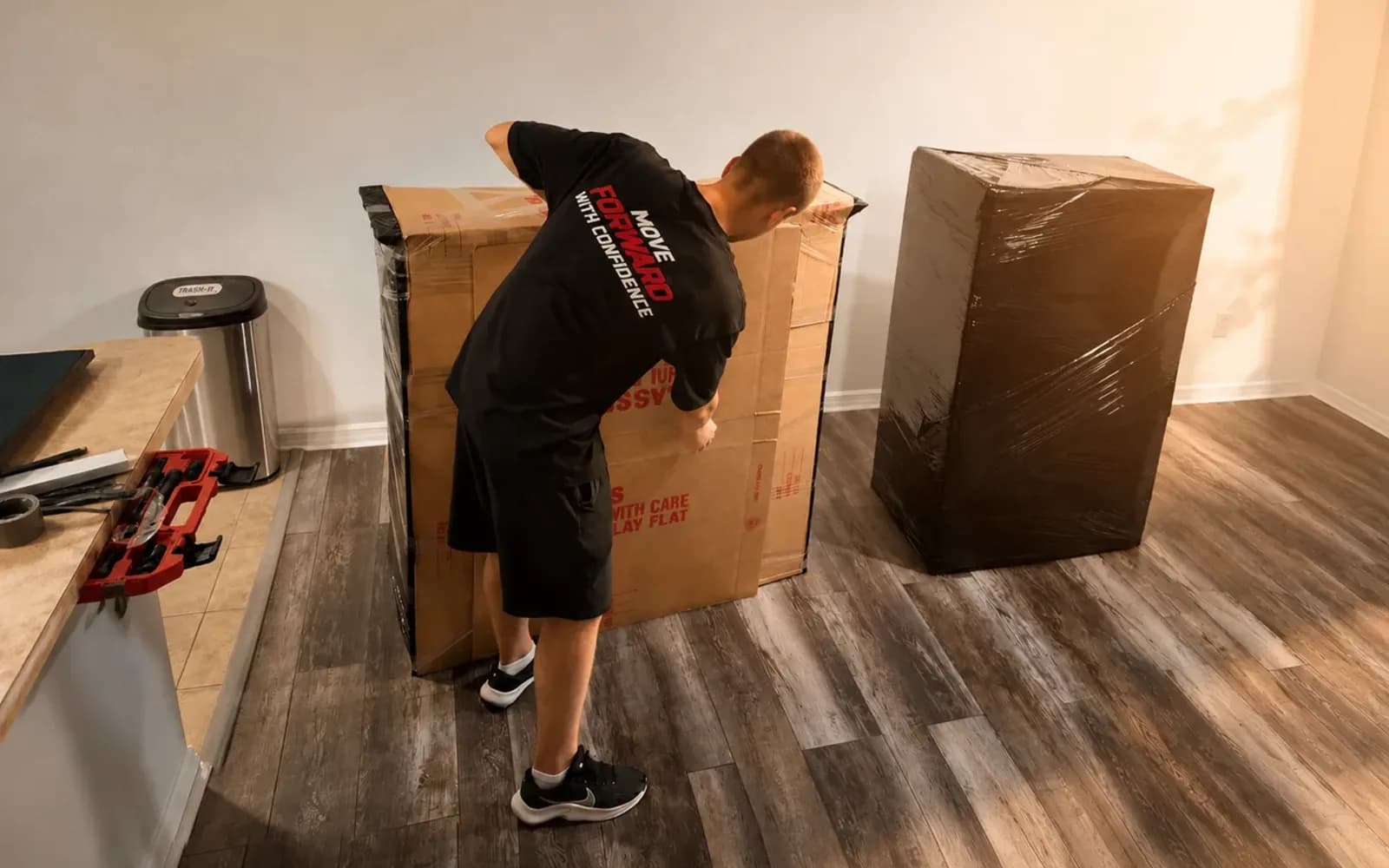 Mover in a black shirt sealing a large carton beside wrapped furniture in a living room