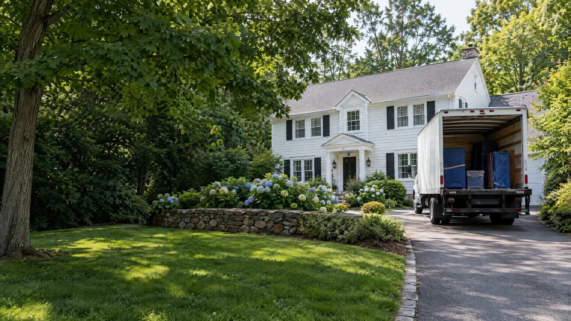 White moving truck in the driveway of a leafy New England colonial home with a broad green lawn