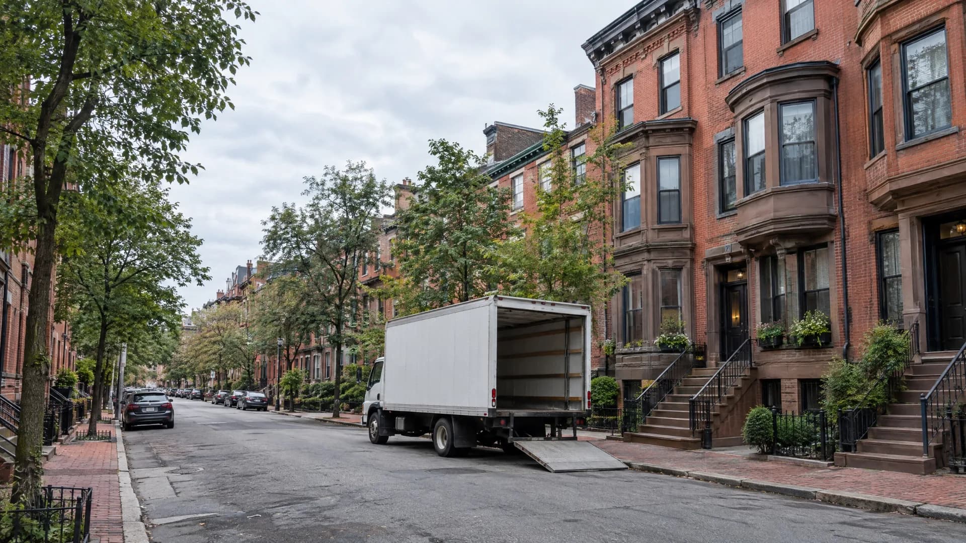 White moving truck parked on a tree-lined Boston residential block with red-brick rowhouses and brownstone stoops