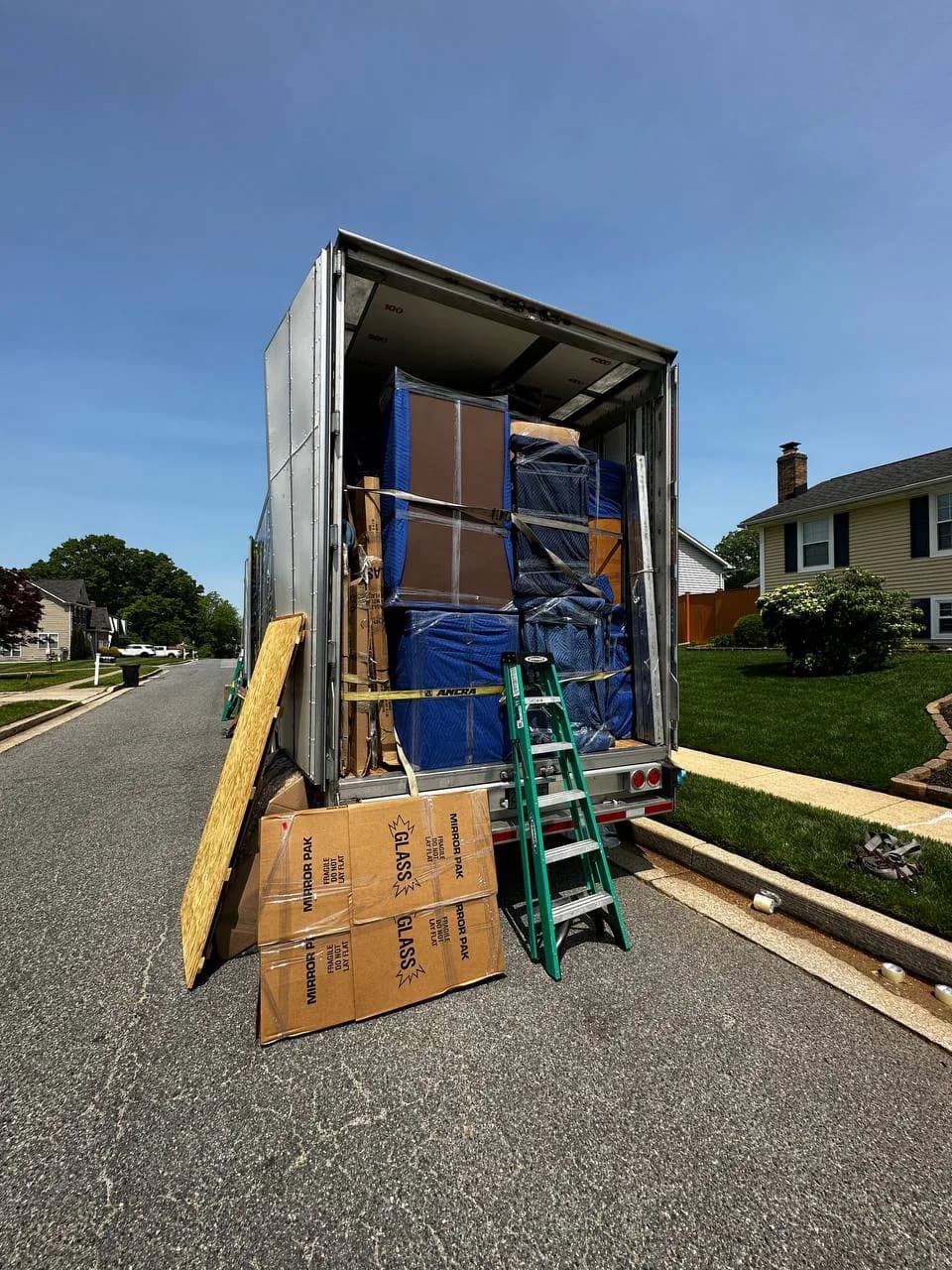 An open moving trailer packed with pad-wrapped furniture, parked on a residential street outside a house