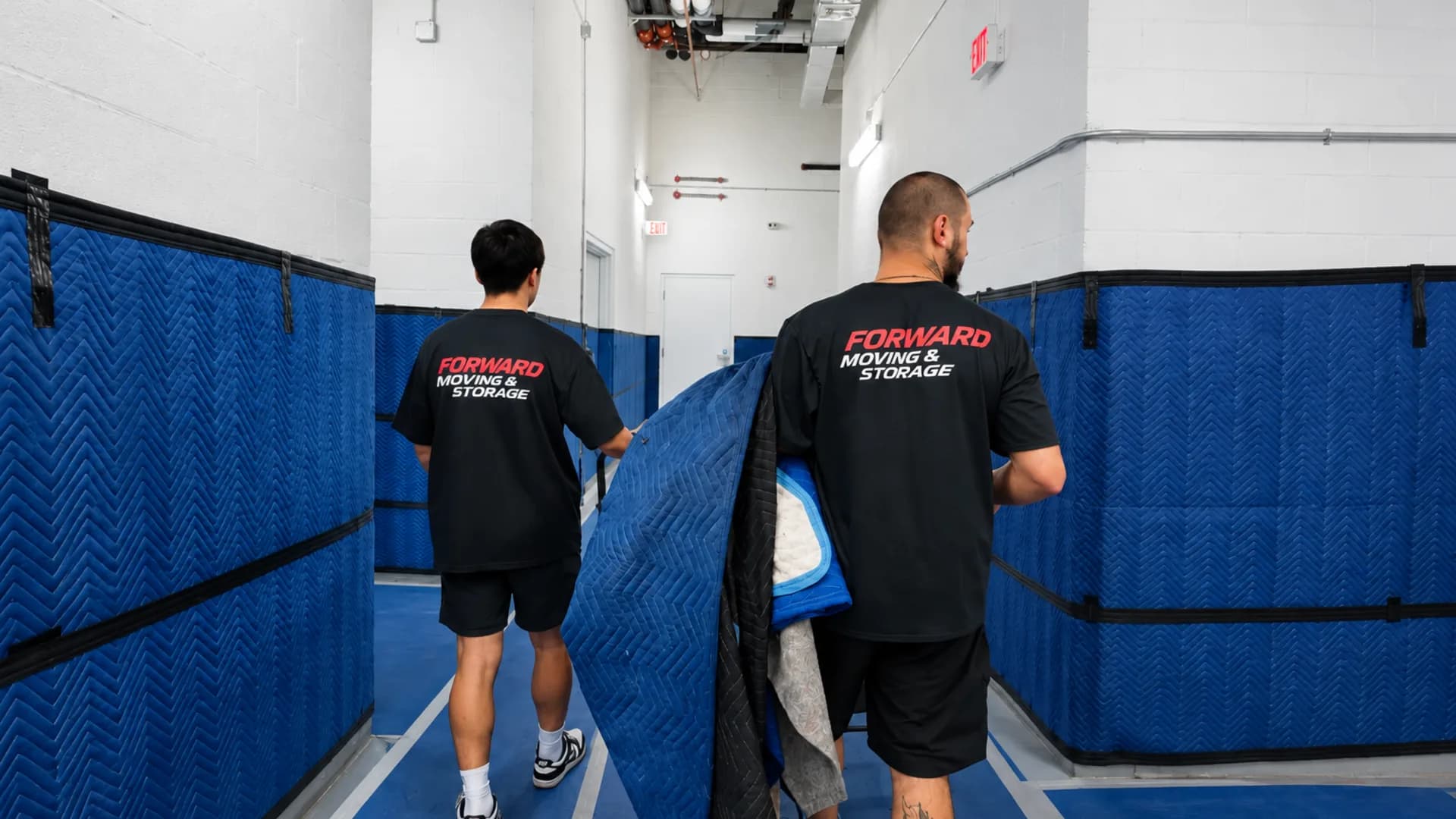 Two movers in branded shirts carrying moving blankets down a building corridor