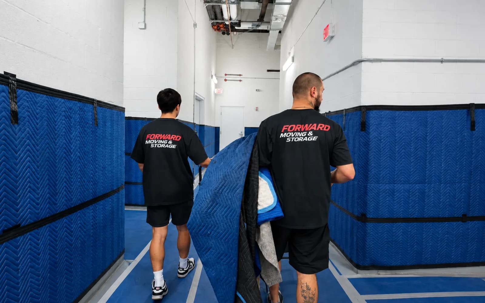 Two movers in Forward shirts carrying moving blankets down a building service corridor lined with protective padding