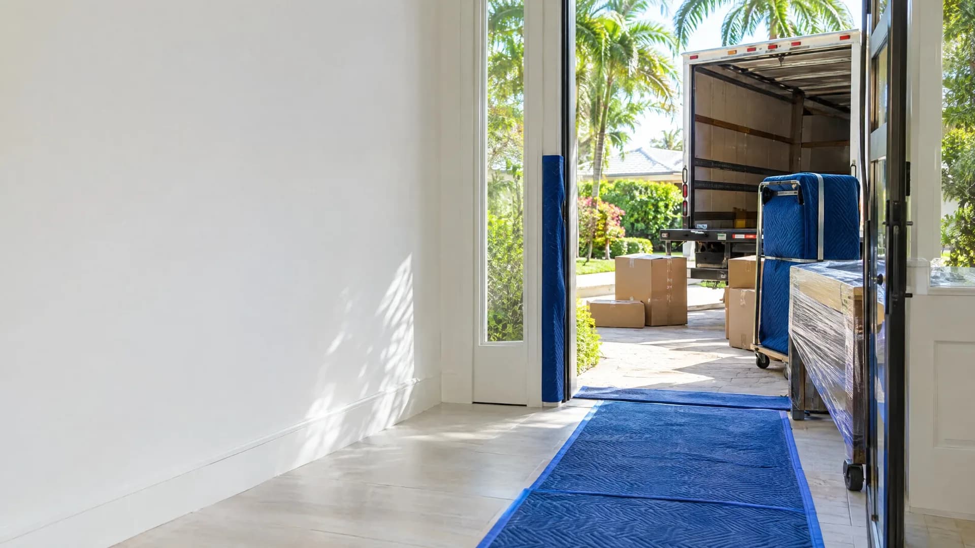 Bright Florida entryway with blue floor protection, wrapped furniture, moving boxes, and an open moving truck outside