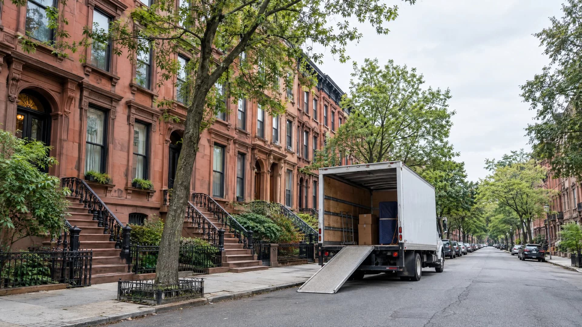 White moving truck loading on a tree-lined New York City brownstone street with brick rowhouses